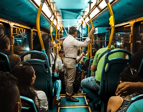 Interior of a crowded city bus with passengers seated and a man standing in the aisle holding a grab rail.