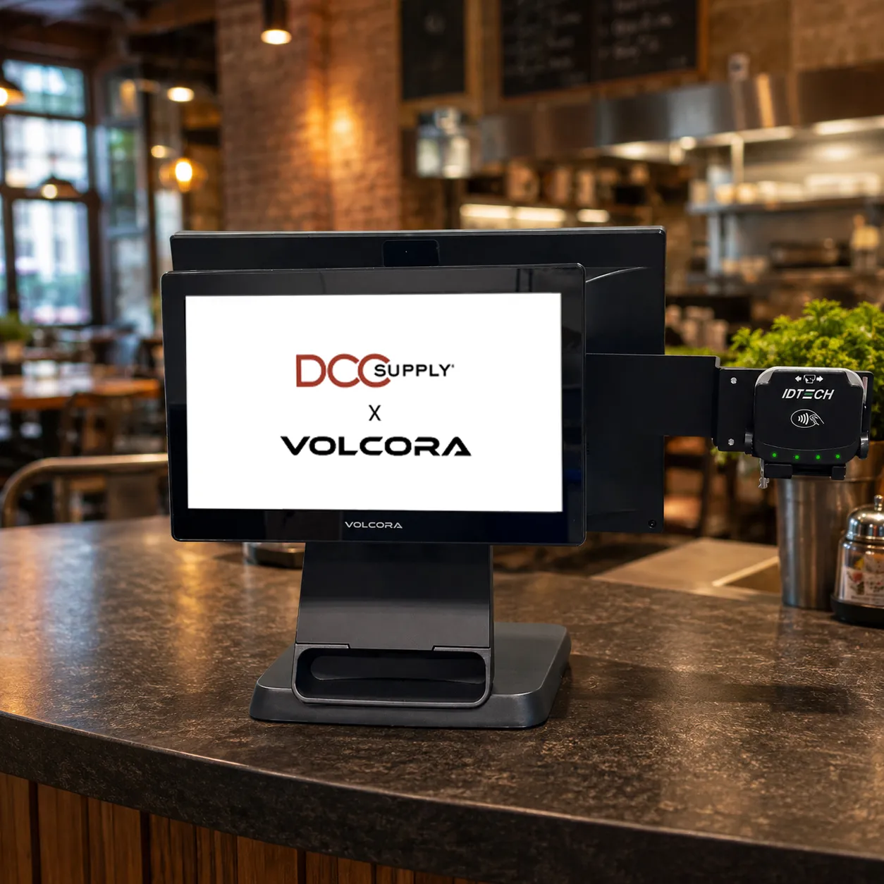 Black Volcora POS terminal on a cafe counter with an ID TECH contactless payment reader mounted on a side arm, screen showing DCC Supply and Volcora logos.