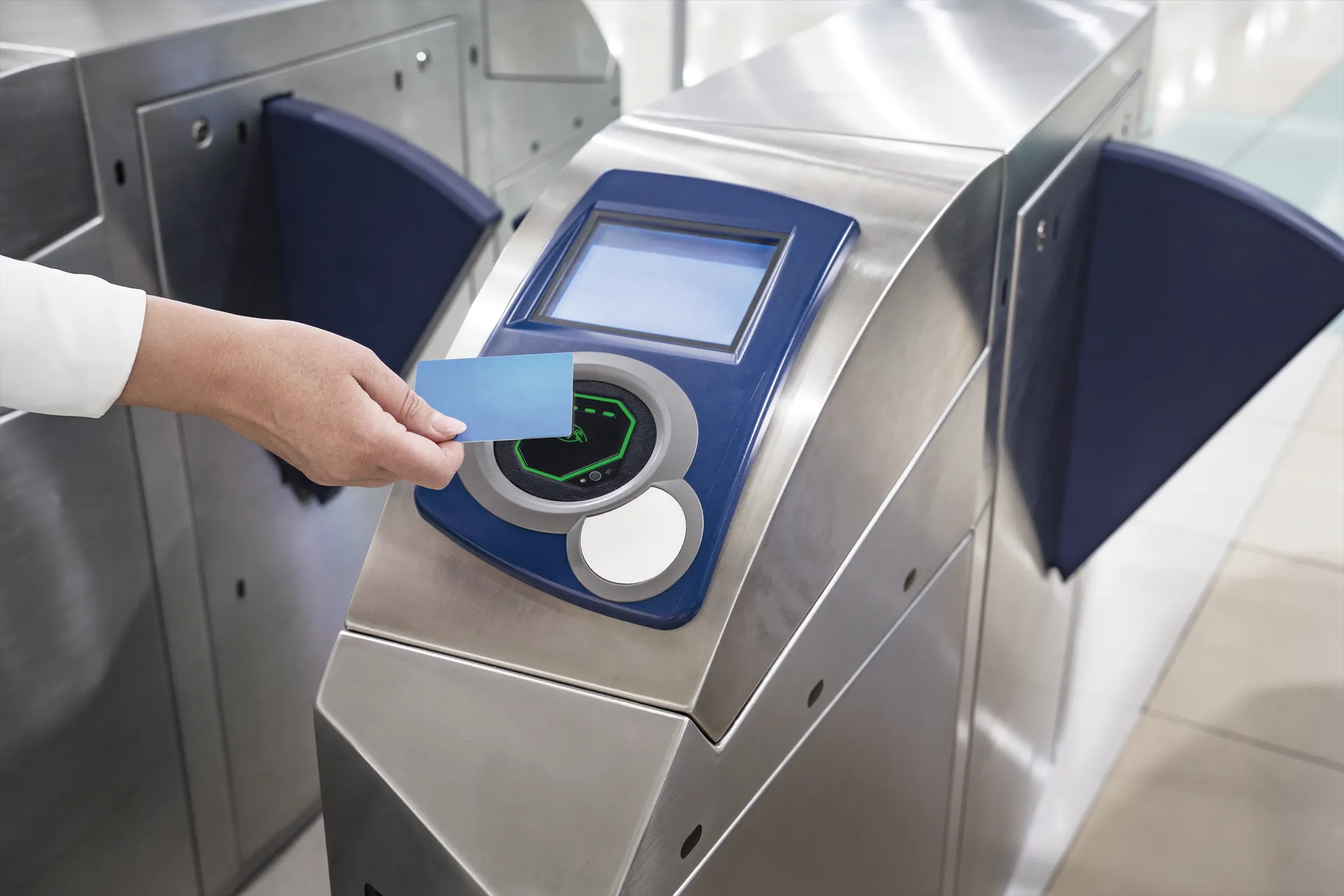 Hand holding a blue bank card over the contactless reader of a stainless steel transit fare gate.